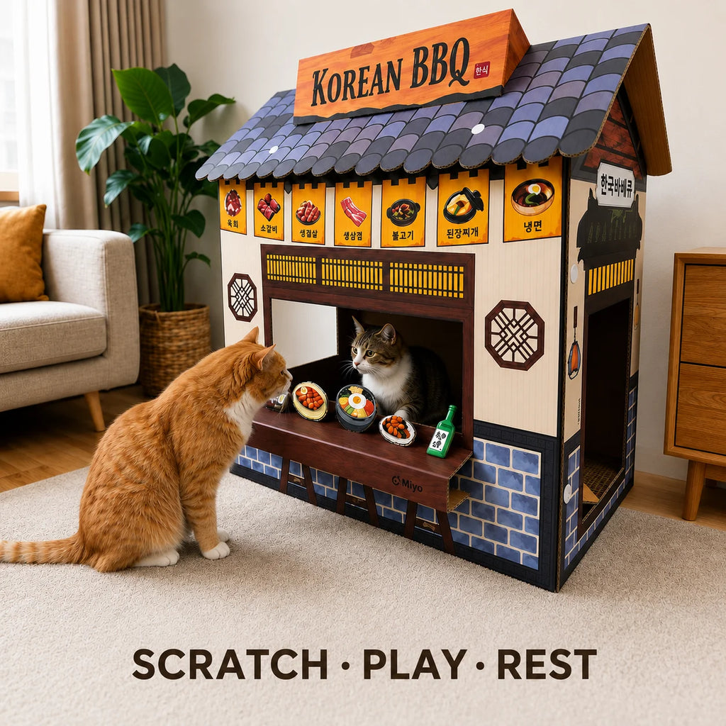 A cat house shaped like a Korean BBQ restaurant, with two cats interacting with it in a living room. The house has a dark gray tiled roof, beige walls, blue brick-patterned base, and a pull-out counter with pretend food bowls and a bottle.