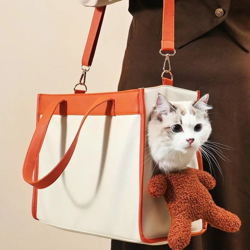 A cream and orange pet carrier tote bag being carried by a person, with a white cat visible through the top opening and a small brown plush toy hanging from the side.
