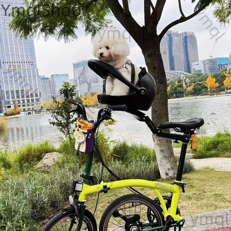 A small white dog sits in a black pet car seat mounted on a bright yellow folding bicycle outdoors by a tree beside a river, with a city skyline in the background.