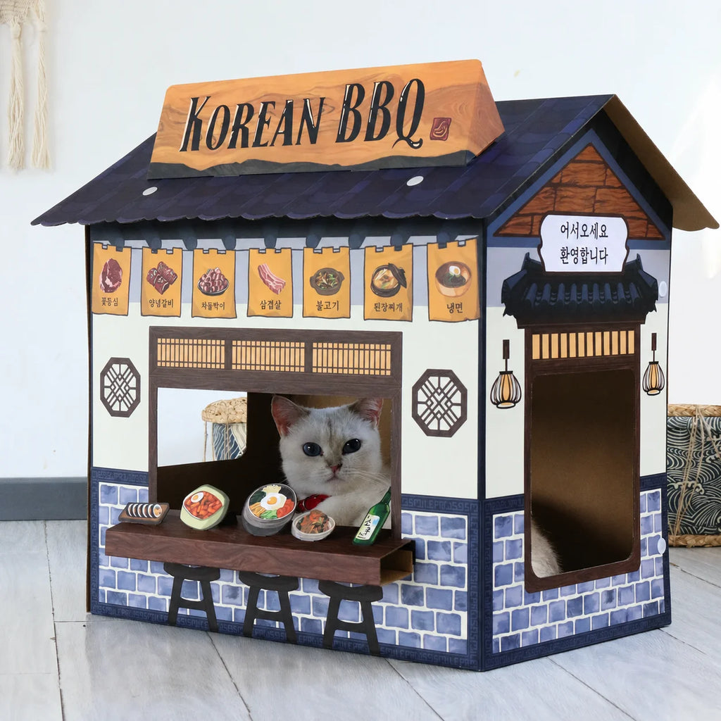 A cat sits inside a miniature house-shaped pet playhouse designed to look like a Korean BBQ restaurant, with a sign reading 'KOREAN BBQ' and food illustrations on the front.