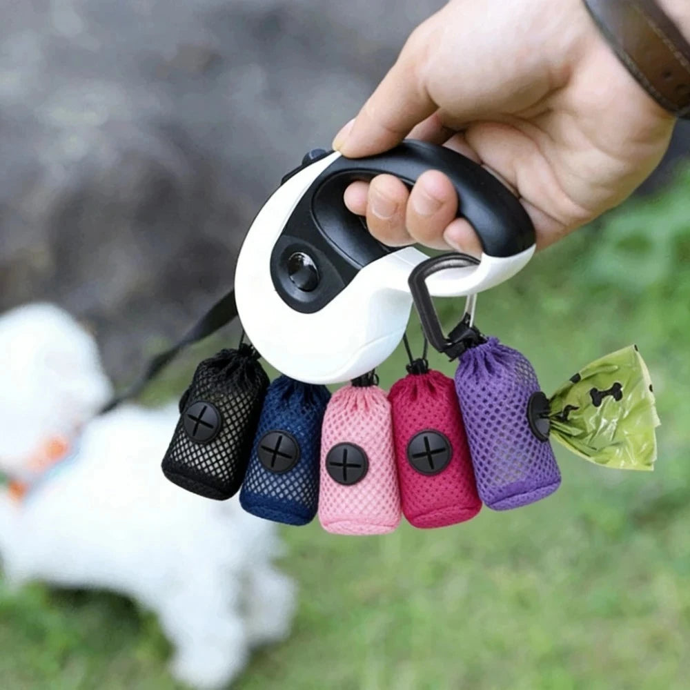 A hand holds a retractable dog leash with four attached mesh poop bags in black, blue, pink, and purple, plus a green plastic bag. The background shows blurred grass and a white dog outdoors.