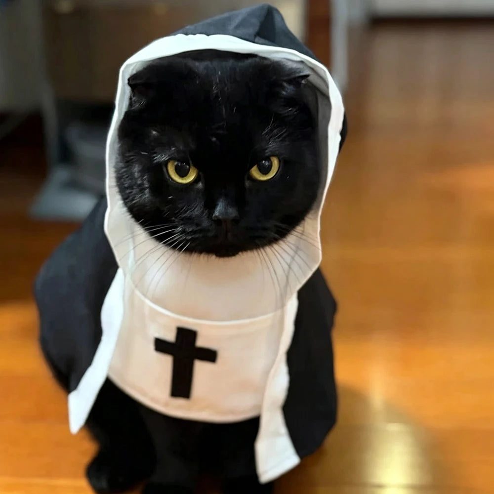A black cat wearing a black and white nun costume with a white cross on the chest, sitting on a wooden floor.