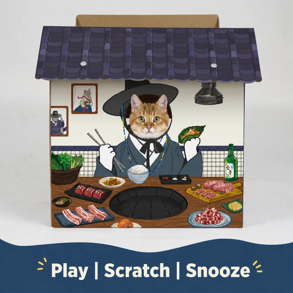 A rectangular cardboard product box with a colorful illustrated scene of a cat dressed in traditional Korean clothing sitting at a table with food, set against a white background with a blue banner at the bottom reading 'Play | Scratch | Snooze'.