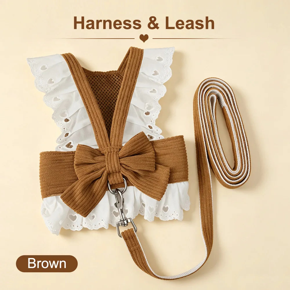 A pet harness and leash set shown on a warm beige background. The harness is brown ribbed fabric with white eyelet lace ruffle trim and a large bow at the front, attached to a matching long leash.