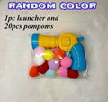 A clear plastic bag containing a yellow and blue toy launcher filled with colorful pom-poms, with text reading 'RANDOM COLOR 1pc launcher and 20pcs pompoms' on a light textured background.