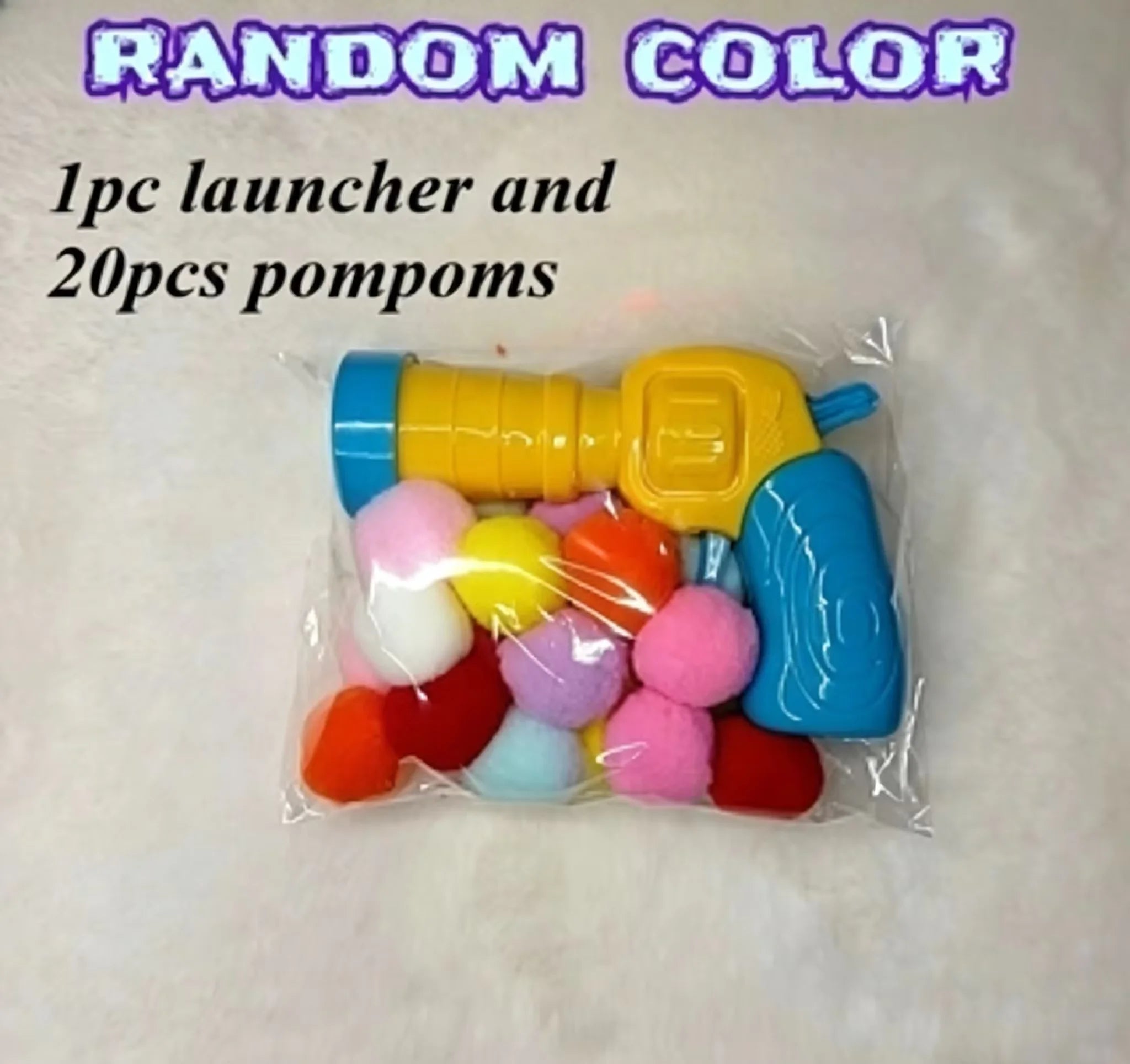 A clear plastic bag containing a yellow and blue toy launcher filled with colorful pom-poms, with text reading 'RANDOM COLOR 1pc launcher and 20pcs pompoms' on a light textured background.