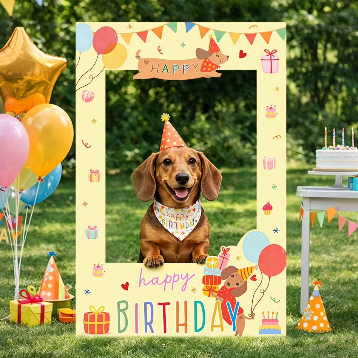 A dachshund wearing a party hat and bandana sits centered inside a large yellow 'Happy Birthday' photo frame backdrop on a grassy lawn, with balloons, gifts, a cake, and party decorations around it.