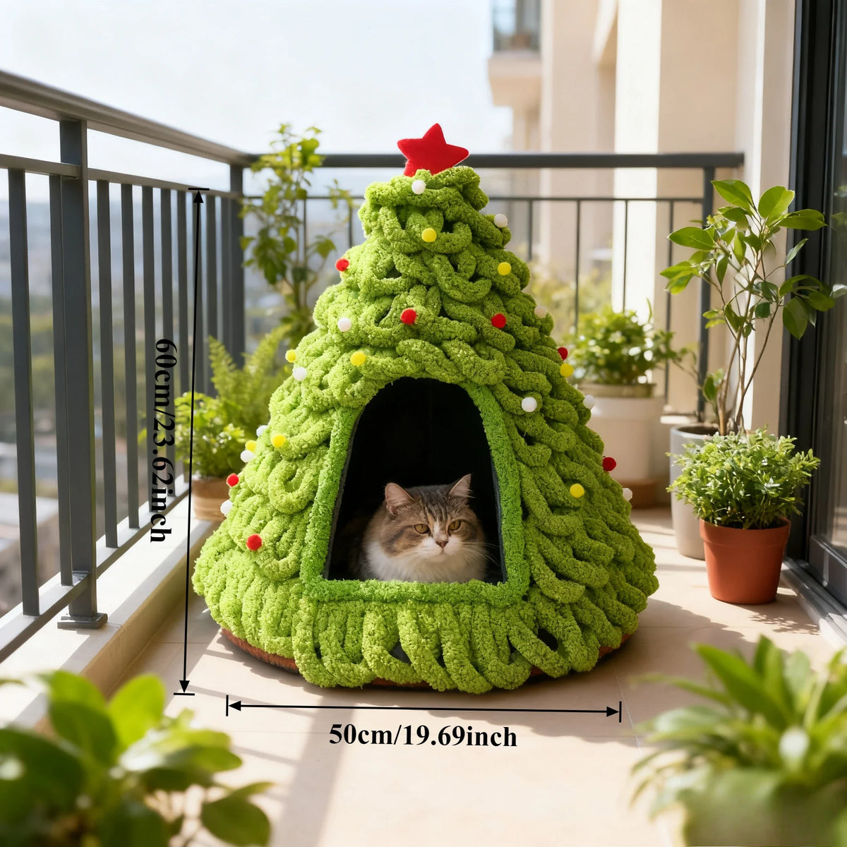 Green knitted cat house shaped like a Christmas tree on a balcony with plants.