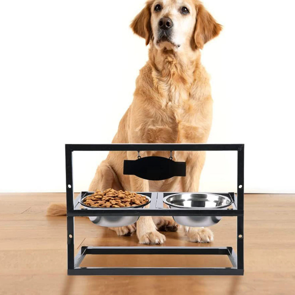 A golden retriever sits behind a black metal elevated pet feeder stand with two stainless steel bowls, one containing dry dog food, on a light wood floor against a white background.