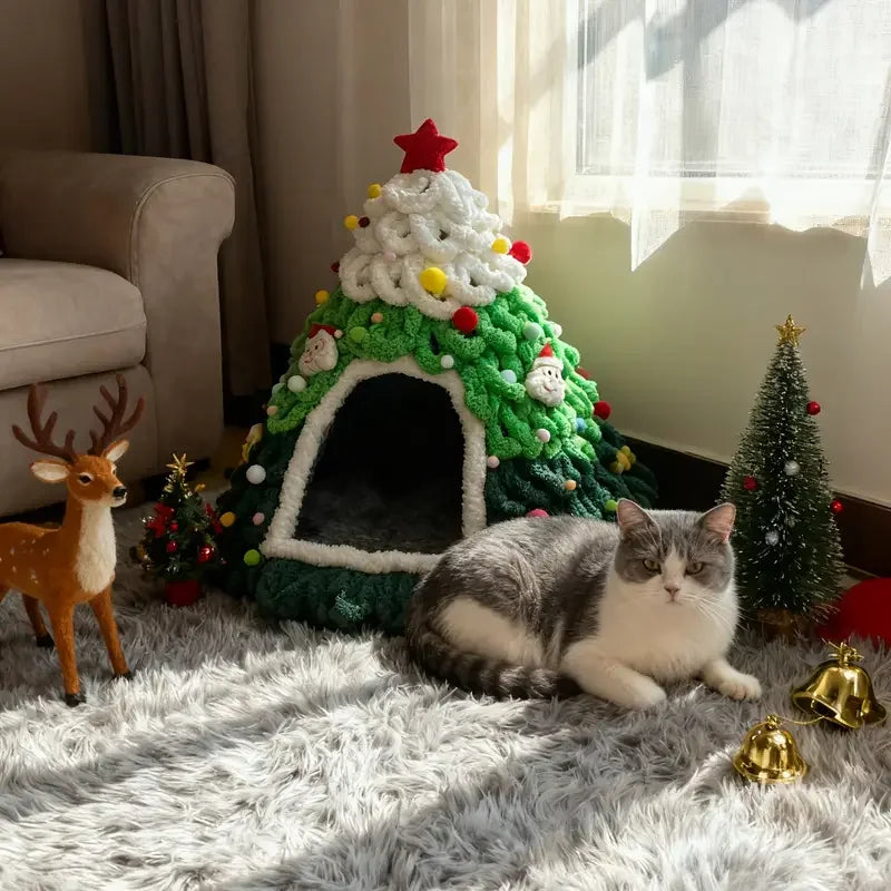 A gray and white cat lies on a fluffy white rug in front of a crocheted Christmas tree-shaped cat bed decorated with a red star, white fluffy trim, and colorful ornaments. The scene is styled like a cozy holiday living room with a small deer figurine, miniature Christmas tree, gold bells, and a sofa in the background.