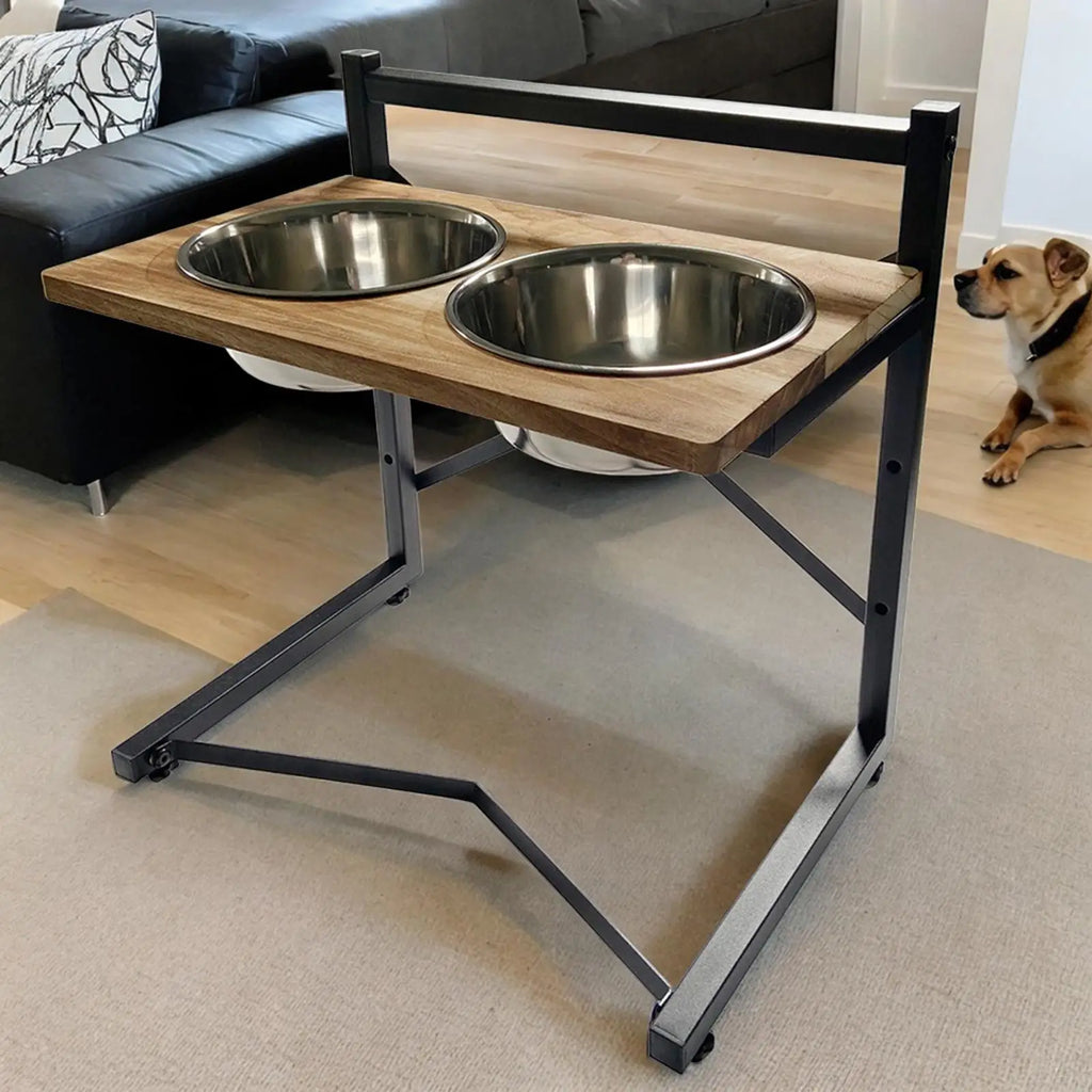 A two-bowl elevated pet feeder and water table with a light wood top and black metal frame, shown in a living room with a dog sitting nearby.