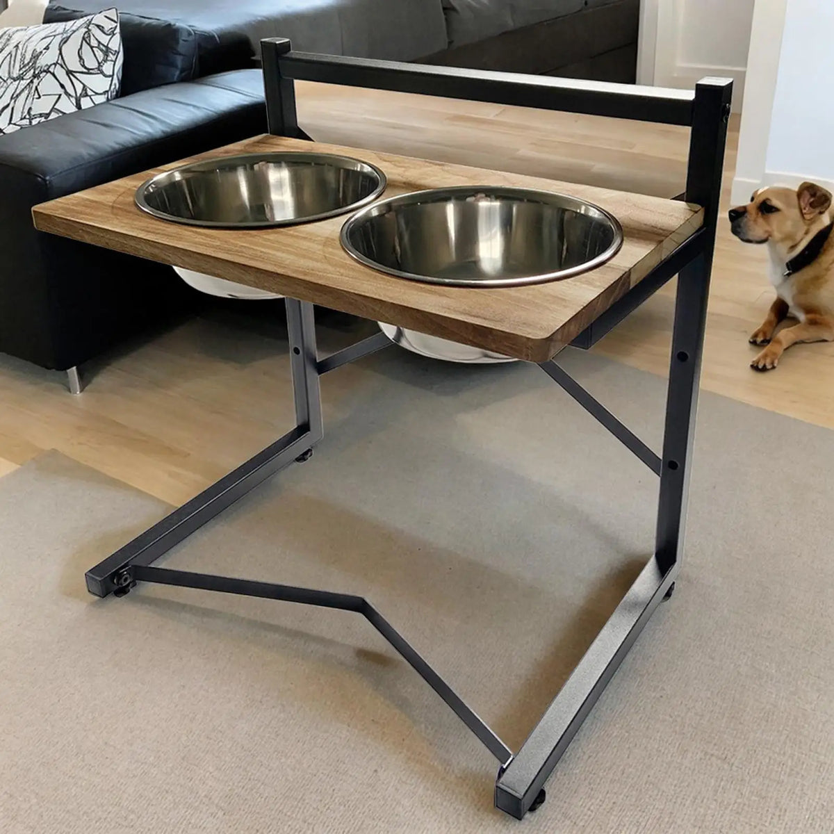 A two-bowl elevated pet feeder and water table with a light wood top and black metal frame, shown in a living room with a dog sitting nearby.