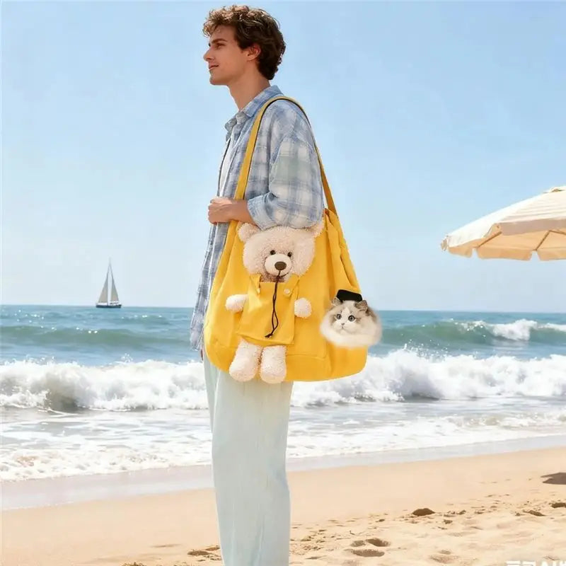 A man stands on a sandy beach wearing a light blue plaid shirt and light pants, carrying a bright yellow shoulder bag with a plush teddy bear and a small dog toy attached. The ocean, blue sky, and a beach umbrella are visible in the background.