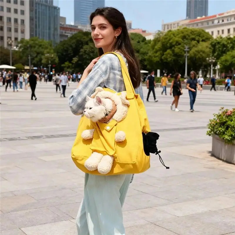 A woman in a city square carries a bright yellow shoulder bag with a plush cream-colored stuffed animal attached to the front. The bag has a drawstring side pocket with a black pouch inside.