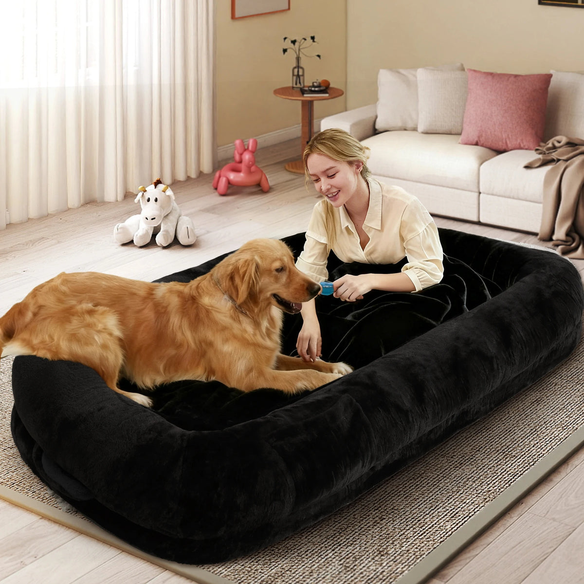 A golden retriever dog rests inside a large black plush pet bed on a rug in a living room, with a woman sitting beside it and holding a blue pet toy.