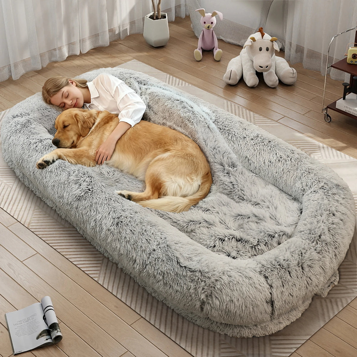A large, plush, fluffy pet bed in light gray is shown in a cozy room setting with a woman and a golden retriever dog resting inside it. The scene includes a wooden floor, a patterned rug, and some plush toys in the background.