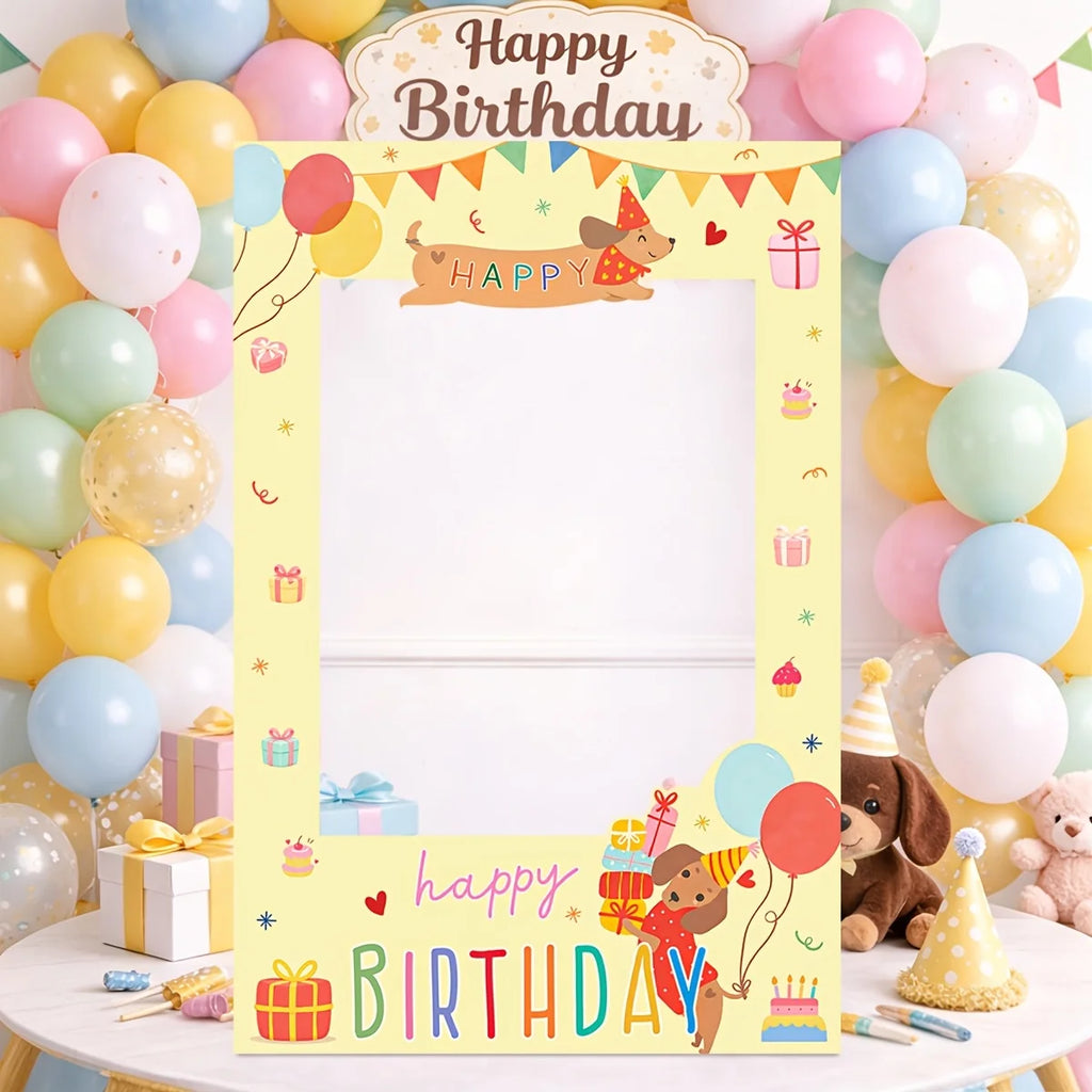 A pastel yellow birthday photo frame with cartoon dog illustrations, balloons, gifts, and colorful 'Happy Birthday' text, displayed on a table with a matching balloon backdrop and party decorations.