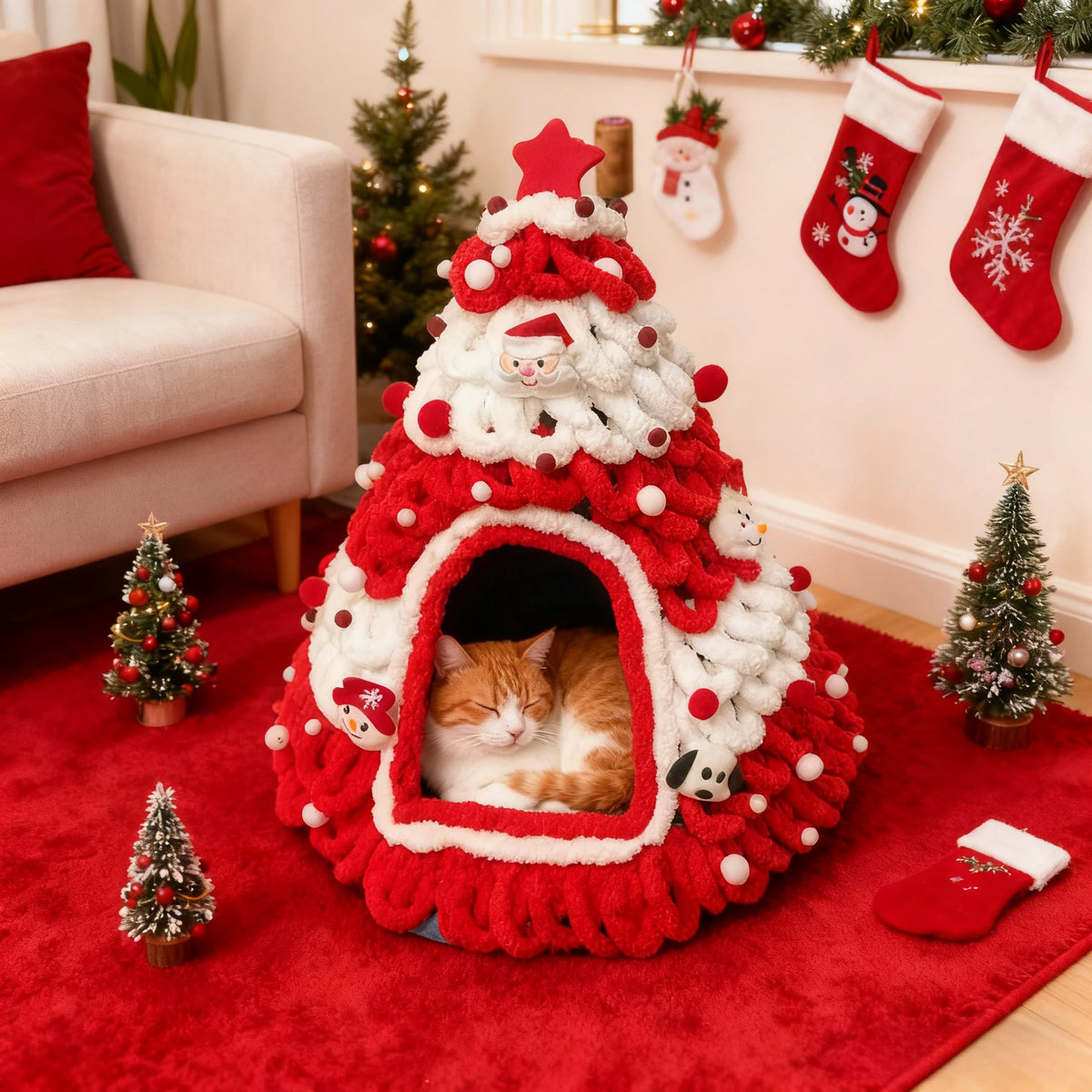 A fluffy red and white Christmas tree-shaped cat bed with a sleeping orange tabby cat inside, styled in a cozy holiday living room with miniature Christmas trees, stockings, and a red rug.