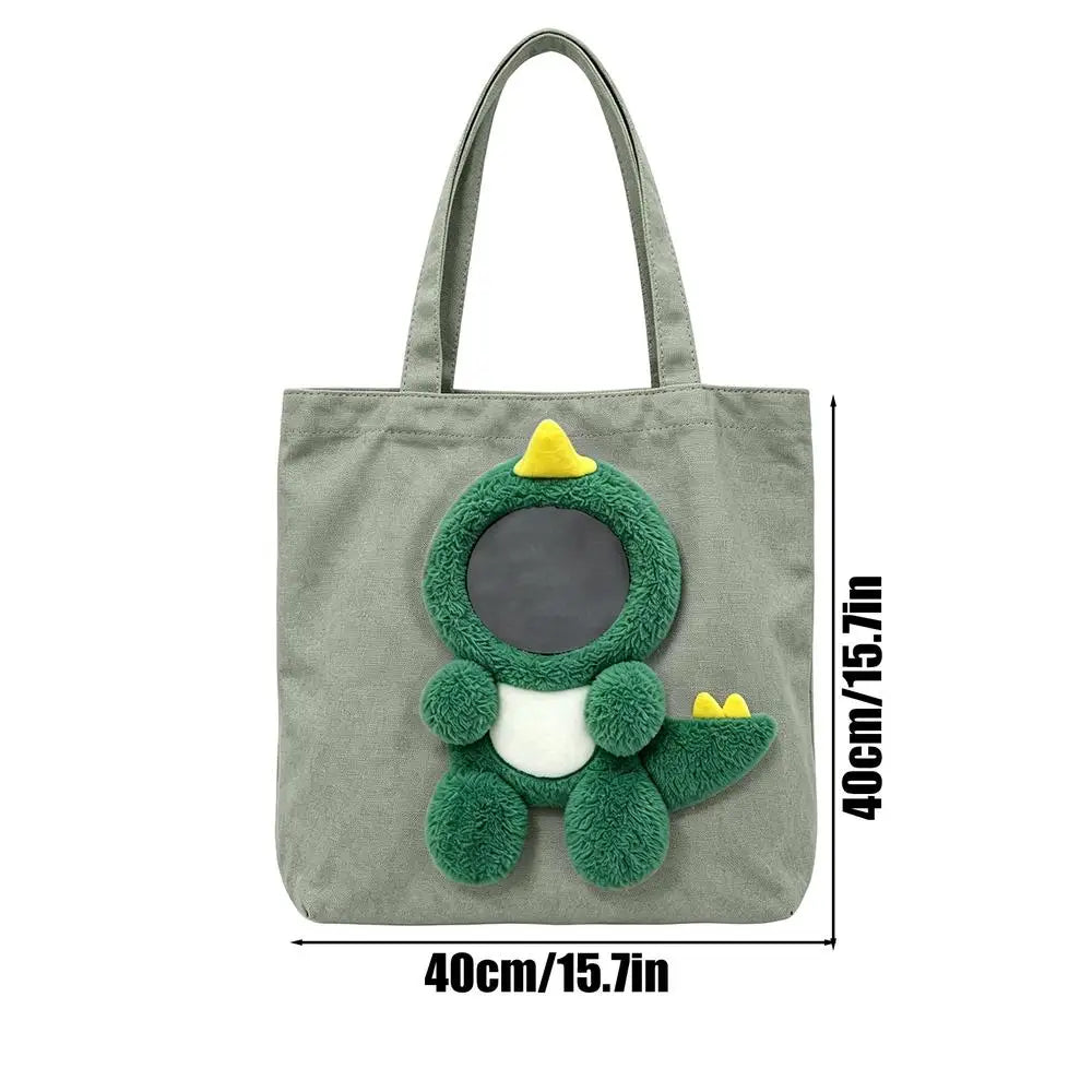 A sage green canvas tote bag with a plush green dinosaur character on the front, featuring a dark circular window and yellow accents. Black lines and text indicate dimensions of 40cm/15.