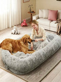 A golden retriever dog rests inside a large, fluffy light gray pet bed while a woman sits beside it on a rug in a cozy living room setting.
