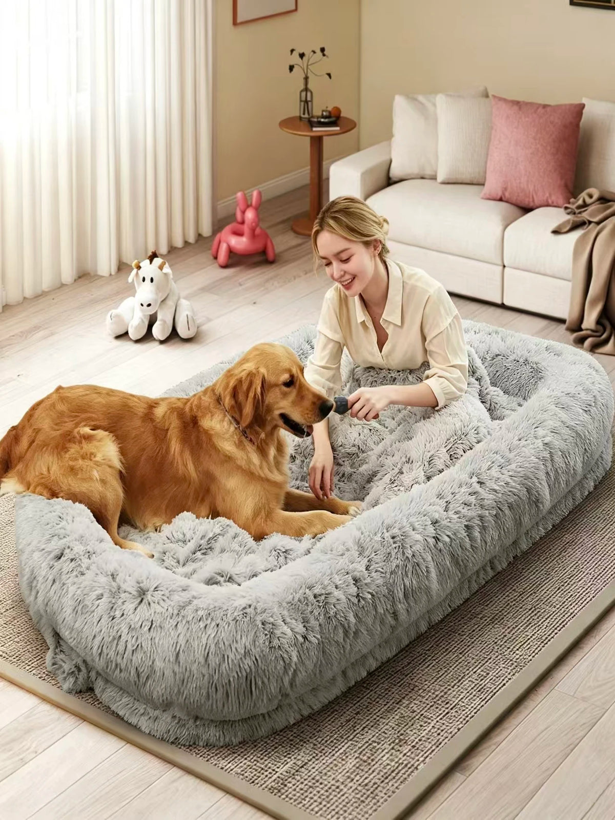 A golden retriever dog rests inside a large, fluffy light gray pet bed while a woman sits beside it on a rug in a cozy living room setting.