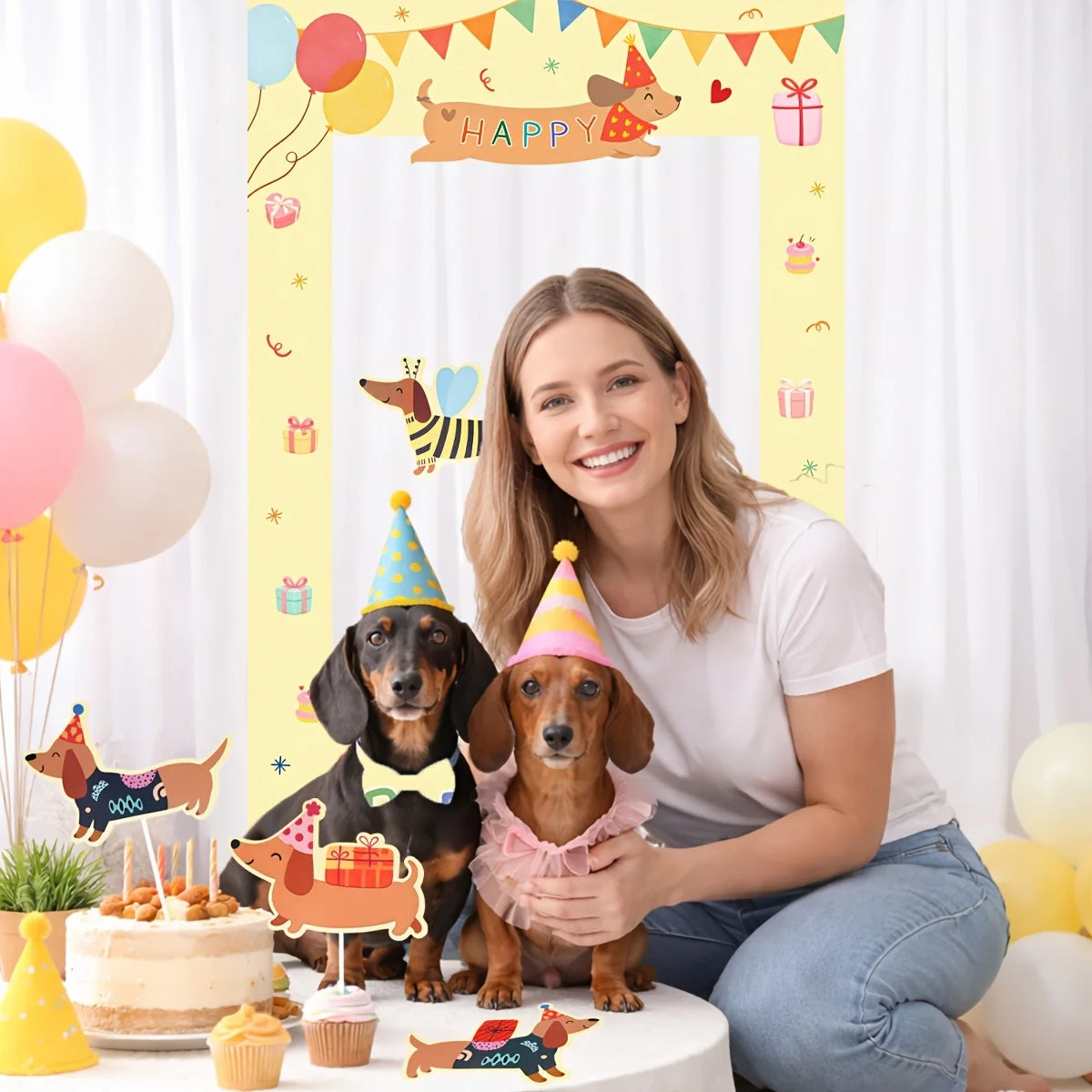 A woman kneels beside two dachshunds wearing party hats in a decorated birthday scene with a large yellow 'Happy Birthday' backdrop, balloons, cupcakes, and dog-shaped cake toppers.
