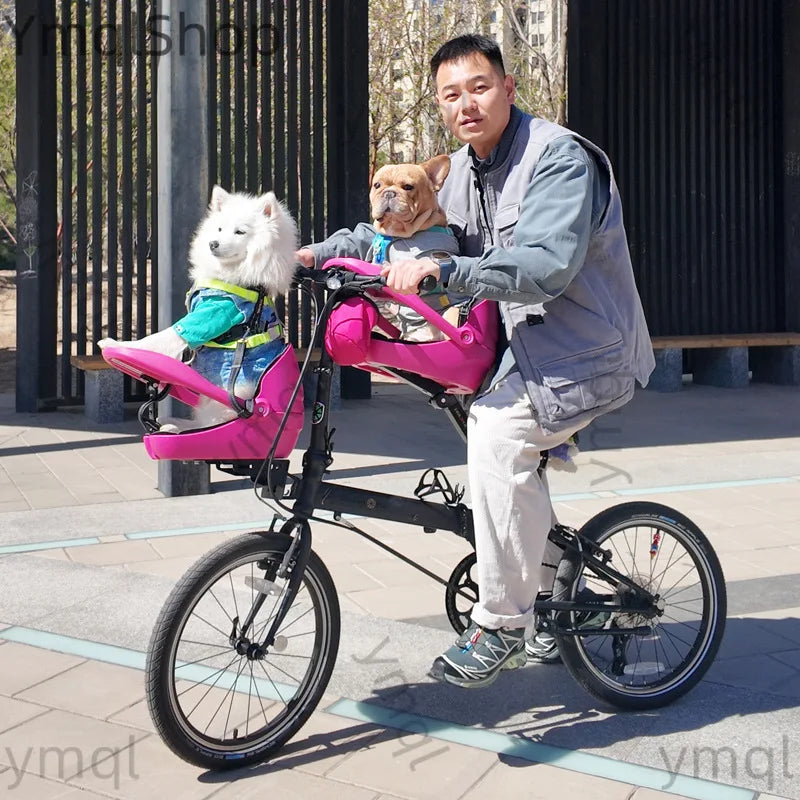 A man rides a black folding bicycle in an outdoor paved area while wearing a bright pink pet bike seat belt harness. A white fluffy dog sits in the front pink pet seat, and a small tan dog sits on the rider's lap.