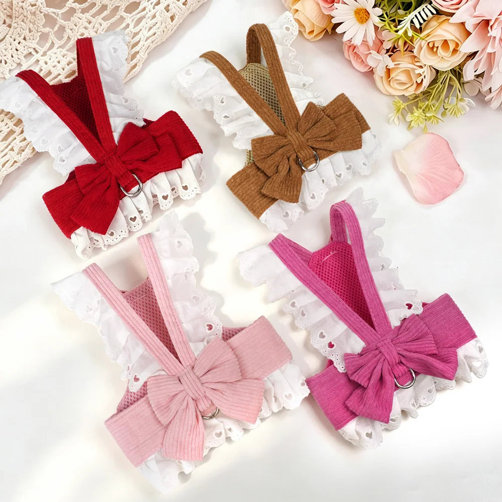 Four pet harnesses arranged on a white surface, each with a large bow at the center, lace trim, and a mesh back. The harnesses come in red, brown, light pink, and magenta pink, with decorative flowers in the top right corner.