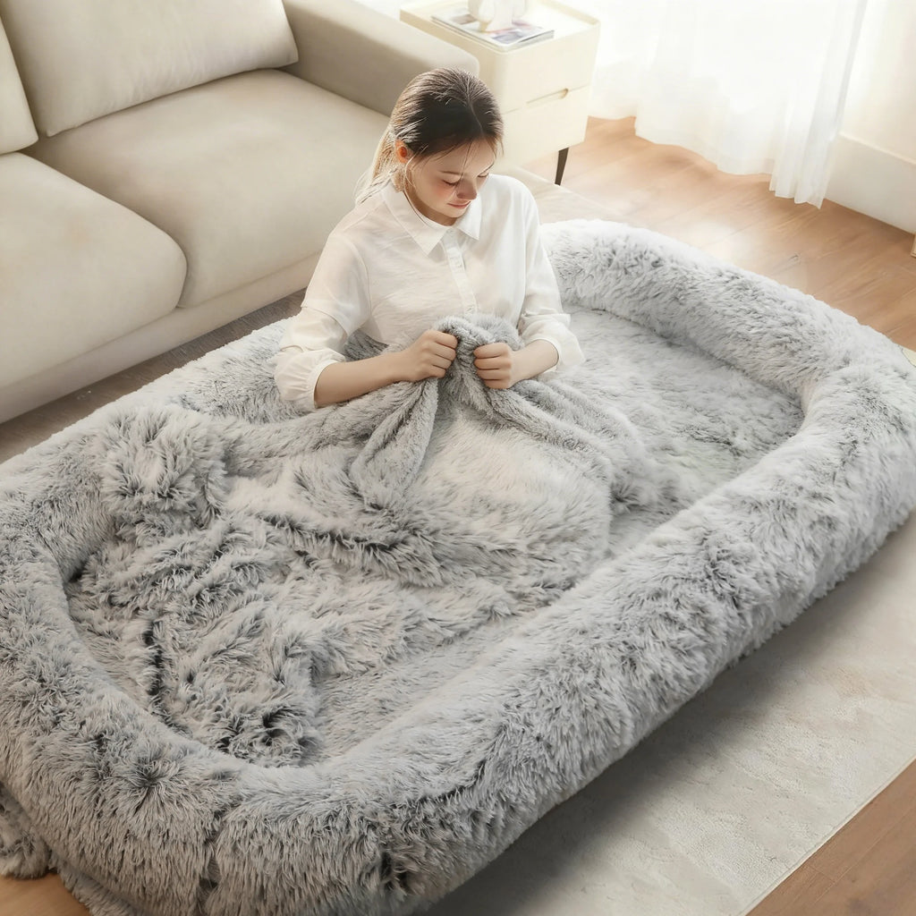 A woman sits inside a large, fluffy gray pet bed or lounge mat in a bright living room, wrapped in a matching gray faux-fur throw blanket.