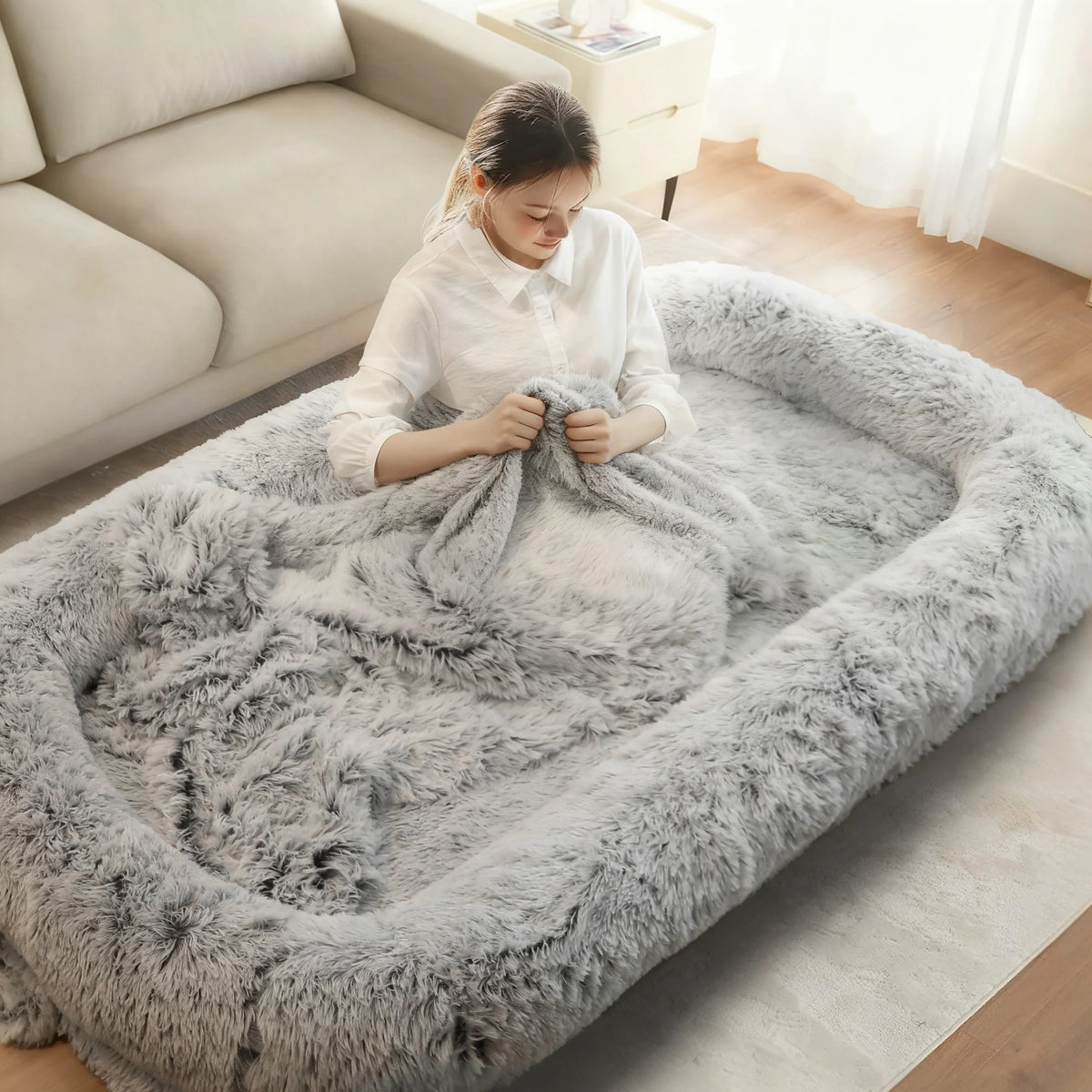 A woman sits inside a large, fluffy gray pet bed or lounge mat in a bright living room, wrapped in a matching gray faux-fur throw blanket.