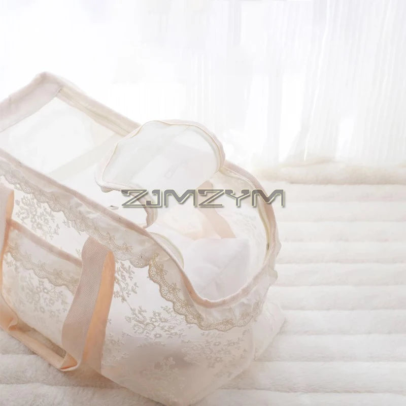 A cream-colored mesh baby bassinet with delicate lace trim and soft fabric handles, shown on a white quilted bed with sheer curtains in the background.